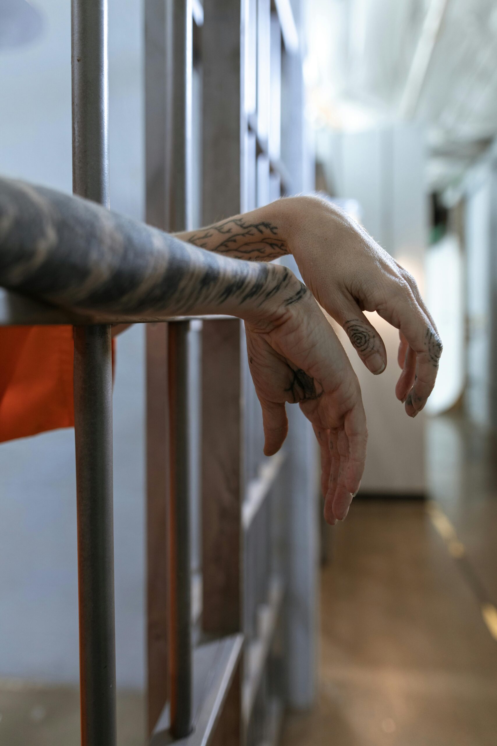 Close-up view of tattooed hands hanging behind prison cell bars, evoking confinement.